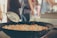A close-up view of a large pan filled with oil where pieces of food are being deep-fried. A skimmer is held above the pan, allowing excess oil to drip back into it. In the background, blurred figures of people suggest a busy environment, possibly a market or festival.