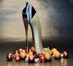A decorative high-heeled shoe shaped like a stiletto with a glossy black upper and golden glittery heel is surrounded by an arrangement of glittering bronze and gold Christmas baubles on a dark, reflective surface.