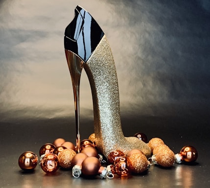 A decorative high-heeled shoe shaped like a stiletto with a glossy black upper and golden glittery heel is surrounded by an arrangement of glittering bronze and gold Christmas baubles on a dark, reflective surface.