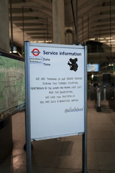 A service information board at a subway station displays a message of solidarity with Australia, expressing sorrow for the loss of life and devastation. The board includes social media handles and a shadow-like illustration of a koala.