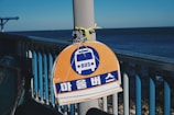 A bus stop sign with Korean characters is attached to a pole on a railing by the seaside, with a clear view of the ocean in the background under a blue sky.