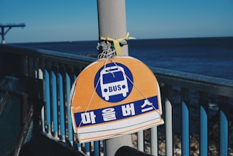 A bus stop sign with Korean characters is attached to a pole on a railing by the seaside, with a clear view of the ocean in the background under a blue sky.