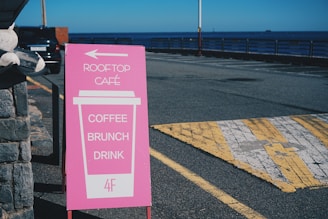 A pink signboard stands on a street next to a stone wall. It has a graphic of a large coffee cup with the words 'ROOFTOP CAFÉ,' 'COFFEE,' 'BRUNCH,' and 'DRINK' printed on it. An arrow points left, and '4F' is indicated at the bottom. In the background, there is a view of a parking lot, a railing, and the sea under a clear blue sky.
