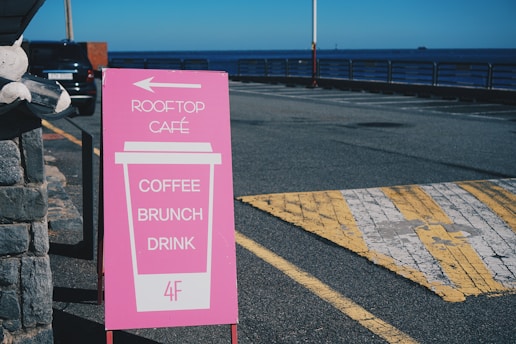 A pink signboard stands on a street next to a stone wall. It has a graphic of a large coffee cup with the words 'ROOFTOP CAFÉ,' 'COFFEE,' 'BRUNCH,' and 'DRINK' printed on it. An arrow points left, and '4F' is indicated at the bottom. In the background, there is a view of a parking lot, a railing, and the sea under a clear blue sky.