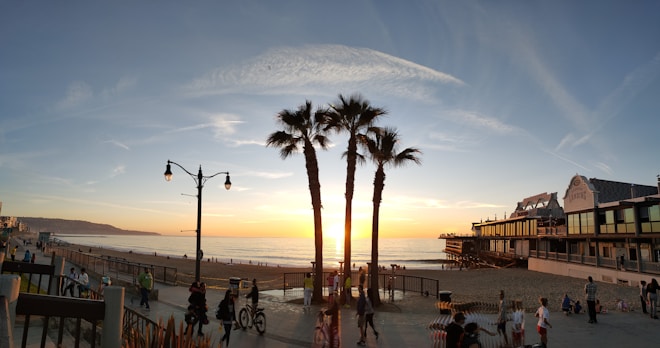 A picturesque beach scene at sunset with the sun setting over the ocean. Palm trees are silhouetted against the vibrant sky, while people stroll along a beachside promenade. A vintage-style street lamp adds charm, and a large building with classic architecture lines the right side of the image.