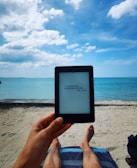 A person relaxes on a sandy beach while holding an e-reader. The clear blue ocean and a partially cloudy sky create a serene backdrop. The person's legs are visible, resting on a beach chair covered with a towel. The scene conveys a sense of leisure and tranquility.