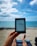 A person relaxes on a sandy beach while holding an e-reader. The clear blue ocean and a partially cloudy sky create a serene backdrop. The person's legs are visible, resting on a beach chair covered with a towel. The scene conveys a sense of leisure and tranquility.