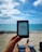 A person relaxes on a sandy beach while holding an e-reader. The clear blue ocean and a partially cloudy sky create a serene backdrop. The person's legs are visible, resting on a beach chair covered with a towel. The scene conveys a sense of leisure and tranquility.