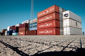 A large stack of colorful shipping containers is arranged neatly on an expansive paved area under a clear blue sky. The containers are primarily red, white, and blue, each labeled with various shipping company logos and identification marks.