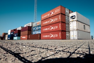 A large stack of colorful shipping containers is arranged neatly on an expansive paved area under a clear blue sky. The containers are primarily red, white, and blue, each labeled with various shipping company logos and identification marks.