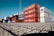 A large stack of colorful shipping containers is arranged neatly on an expansive paved area under a clear blue sky. The containers are primarily red, white, and blue, each labeled with various shipping company logos and identification marks.