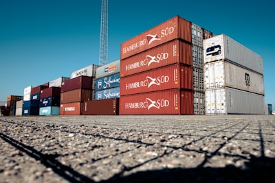 A large stack of colorful shipping containers is arranged neatly on an expansive paved area under a clear blue sky. The containers are primarily red, white, and blue, each labeled with various shipping company logos and identification marks.
