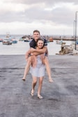 A happy couple is enjoying a playful moment near a waterfront. The woman is carrying the man on her back while both smile broadly. In the background, there are several colorful boats docked at a pier, and a cloudy sky looms above.