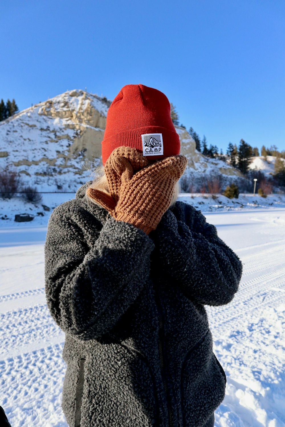 person in red bobble hat, gray sweater, and brown gloves covering face