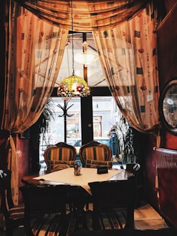 A cozy, intimate café setting with a wooden table and vintage upholstered chairs. Elegant drapes with floral patterns frame a large window overlooking a street, allowing natural light to filter in. A stained glass chandelier hangs from the ceiling, contributing to the warm, inviting atmosphere.