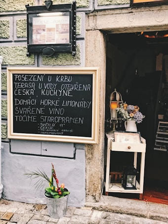 A quaint outdoor setting includes a blackboard with handwritten Czech text advertising menu items like homemade lemonade and mulled wine. Next to it, a small decorative stand holds a lamp and flower arrangement. The wall is textured with a geometric pattern and painted in light green and gray tones. A bucket with a small plant is placed on the cobblestone pavement.