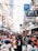 A bustling street market scene crowded with people, showcasing a diversity of storefronts with brightly colored signs and advertisements. Many individuals are browsing or shopping among street vendors offering various goods. The atmosphere is dynamic and lively.