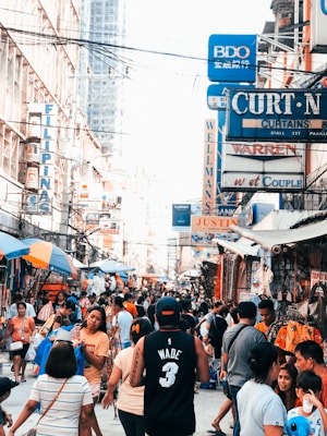 A bustling street market scene crowded with people, showcasing a diversity of storefronts with brightly colored signs and advertisements. Many individuals are browsing or shopping among street vendors offering various goods. The atmosphere is dynamic and lively.