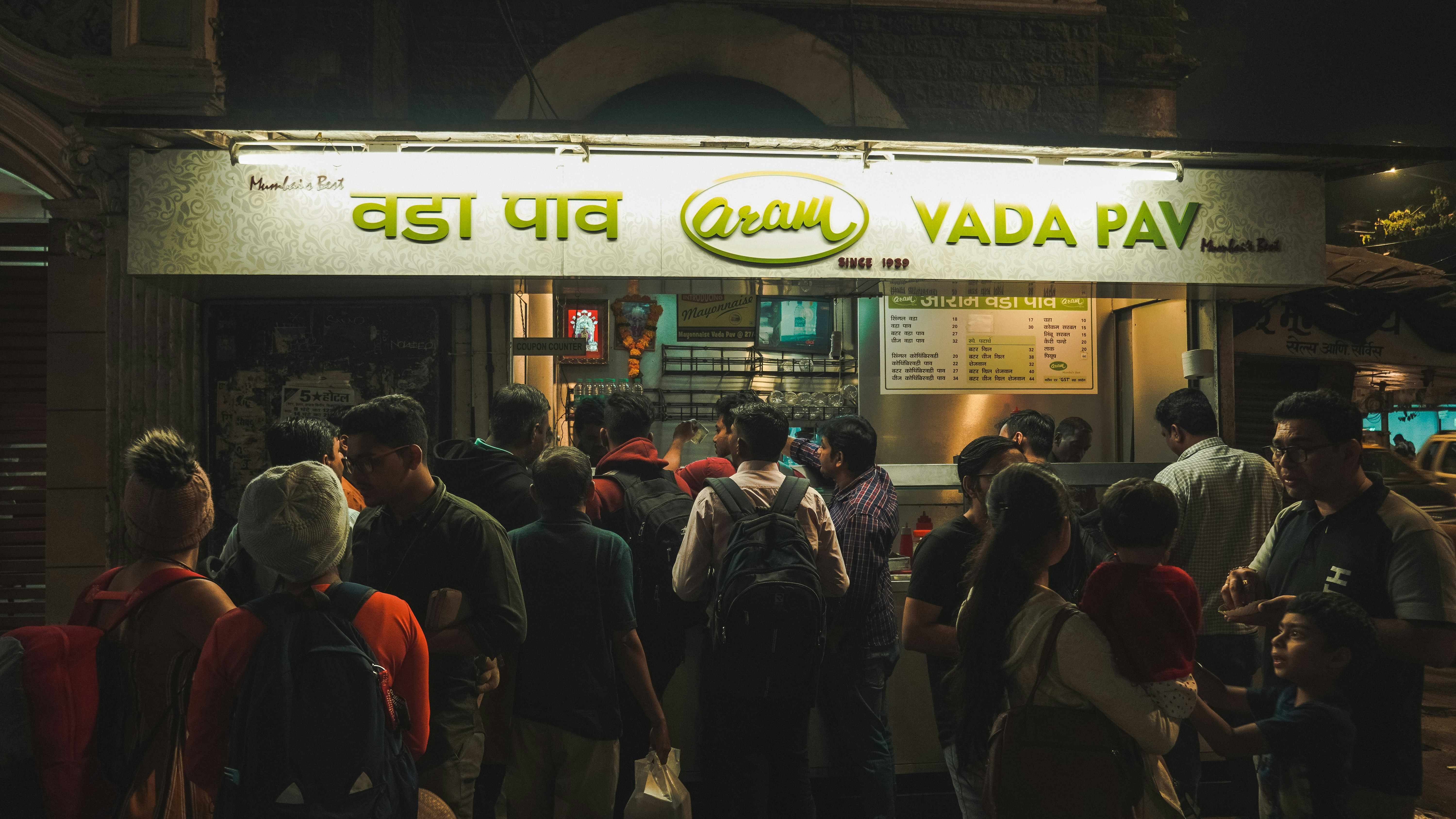 Mumbai street food stall at night, glowing lights, vendors serving vada pav, busy dark street scene, low-key urban lighting