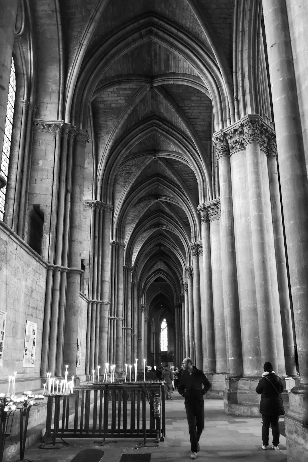 grayscale photo of man walking near candles on stand under arch ceiling