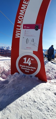 A red and white sign stands on a snow-covered landscape. The sign displays text indicating a 14 km long illuminated toboggan run, and includes a welcome message in German 'WILLKOMMEN'. The background shows snowy mountains and a clear blue sky. Two people stand near a railing, gazing at the landscape.