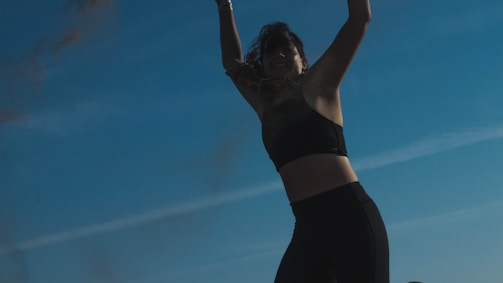 A person is silhouetted against a clear blue sky, appearing to be in motion with arms raised. They are wearing athletic clothing, including a sports bra and leggings.