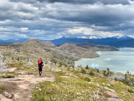 A lone hiker carrying a large red backpack stands on a rocky path, surrounded by a breathtaking landscape of rolling hills and sparse vegetation. In the distance, a vast lake dotted with ice floats and framed by snow-capped mountains stretches under a dramatic cloudy sky.