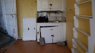A small, worn-out kitchen area features a white sink and countertop with peeling paint. The upper and lower cabinets are closed, with visible damage and missing pieces. There are rocks resting against the cabinets, possibly to keep them closed, and miscellaneous items on the countertop. The walls are a faded yellow with built-in shelving to the right, and the floor is covered with brownish-red tiles.