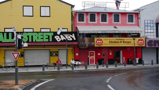 A street scene featuring colorful buildings with various businesses. The left building is painted in yellow with black trim, featuring a sign for baby accessories. Adjacent is a red building with signage for a restaurant and steakhouse. People are sitting and walking along the sidewalk.