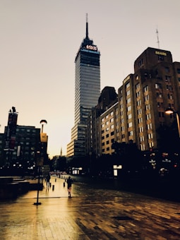A tall skyscraper with a digital clock and the words 'LATINO SEGUROS' at the top, surrounded by other buildings, stands against a clear sky. The ground is wet, reflecting the buildings and streetlights. Pedestrians are walking on the sidewalk.