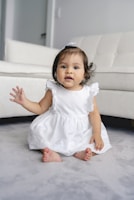 toddler wearing white dress sitting beside white sofa