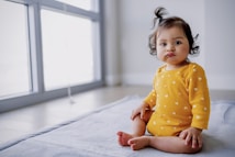 A toddler with dark hair wearing a yellow dress with white heart patterns sits on a light-colored blanket. The child has a small tuft of hair up and an expression of curiosity or contemplation. Natural light from large windows illuminates the room, casting soft shadows.