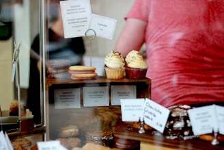 A display of assorted baked goods, featuring cupcakes with sprinkles on a wooden stand, surrounded by various cookies and pastries with price tags. Some of the tags identify items like flourless chocolate pecan sandwich cookies and whoopie pies. A blurred figure is visible in the background.