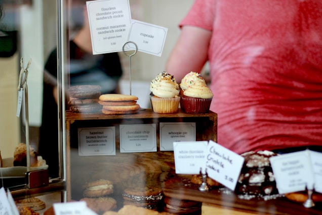 A display of assorted baked goods, featuring cupcakes with sprinkles on a wooden stand, surrounded by various cookies and pastries with price tags. Some of the tags identify items like flourless chocolate pecan sandwich cookies and whoopie pies. A blurred figure is visible in the background.