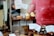 A display of assorted baked goods, featuring cupcakes with sprinkles on a wooden stand, surrounded by various cookies and pastries with price tags. Some of the tags identify items like flourless chocolate pecan sandwich cookies and whoopie pies. A blurred figure is visible in the background.