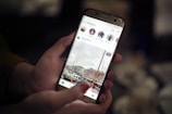 A person's hands holding a smartphone displaying an Instagram feed. The screen shows a photo of a marina with boats and buildings in the background. The focus is on the phone, with a blurred background suggesting an indoor setting with dim lighting.