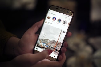 A person's hands holding a smartphone displaying an Instagram feed. The screen shows a photo of a marina with boats and buildings in the background. The focus is on the phone, with a blurred background suggesting an indoor setting with dim lighting.