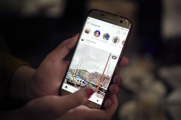 A person's hands holding a smartphone displaying an Instagram feed. The screen shows a photo of a marina with boats and buildings in the background. The focus is on the phone, with a blurred background suggesting an indoor setting with dim lighting.