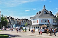 A bustling town square is lined with picturesque, European-style buildings featuring mansard roofs. People, including families and groups, stroll along the cobblestone pathways. Bright signage and flower arrangements along the pathways add to the charm.