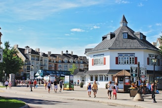 A bustling town square is lined with picturesque, European-style buildings featuring mansard roofs. People, including families and groups, stroll along the cobblestone pathways. Bright signage and flower arrangements along the pathways add to the charm.