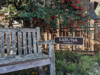 A weathered wooden bench is situated in a garden area surrounded by lush greenery and vibrant red berries. A wooden sign with the words 'KARUNA Compassion' is prominently displayed beside the bench. The background features a wooden fence, with sunlight filtering through the leaves, creating a peaceful and serene atmosphere.