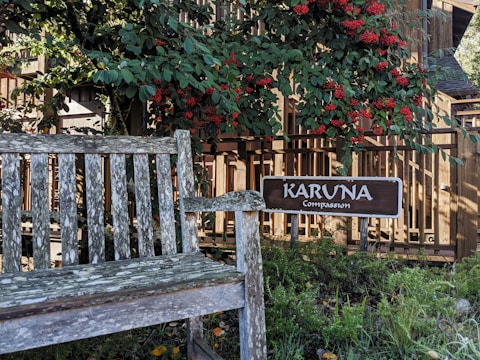 A weathered wooden bench is situated in a garden area surrounded by lush greenery and vibrant red berries. A wooden sign with the words 'KARUNA Compassion' is prominently displayed beside the bench. The background features a wooden fence, with sunlight filtering through the leaves, creating a peaceful and serene atmosphere.