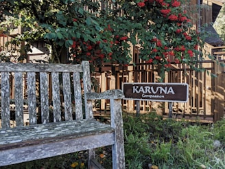 A weathered wooden bench is situated in a garden area surrounded by lush greenery and vibrant red berries. A wooden sign with the words 'KARUNA Compassion' is prominently displayed beside the bench. The background features a wooden fence, with sunlight filtering through the leaves, creating a peaceful and serene atmosphere.