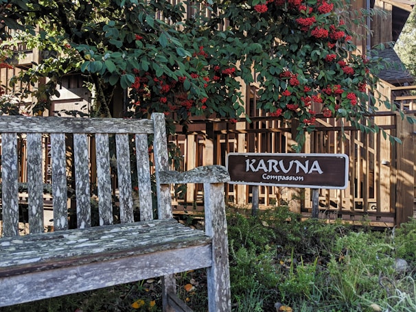 A weathered wooden bench is situated in a garden area surrounded by lush greenery and vibrant red berries. A wooden sign with the words 'KARUNA Compassion' is prominently displayed beside the bench. The background features a wooden fence, with sunlight filtering through the leaves, creating a peaceful and serene atmosphere.