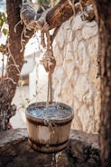 A rustic wooden bucket filled with water hangs from a sturdy, weathered tree branch. Water spills over the edges, creating small streams as it drips down. The scene is set against a stone wall, partially blurred in the background, and surrounded by dry, twisted branches.