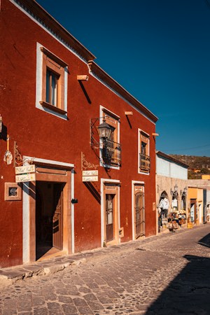 A vibrant street scene featuring a rustic building with a rich terracotta facade and white trim. It has ornate wrought iron balconies and a vintage-style lamp. The cobblestone street adds an old-world charm, and nearby shops display colorful merchandise under a clear blue sky.