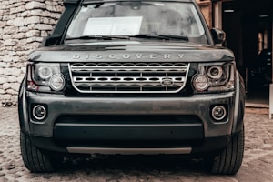 A front view of a Land Rover Discovery vehicle parked on a cobblestone surface. The car's distinctive grille and headlights are prominently visible, with the brand logo and name 'Discovery' clearly displayed above the grille. The image features a rugged stone wall on the left side in the background.
