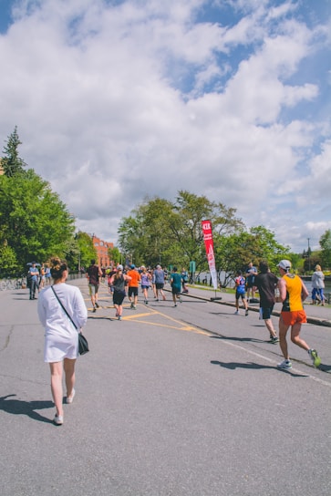 A group of people are participating in an outdoor running event on a sunny day. They are running on a paved road surrounded by lush green trees and under a partly cloudy sky. A red sign with white text is visible in the background.