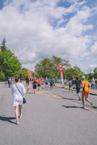 A group of people are participating in an outdoor running event on a sunny day. They are running on a paved road surrounded by lush green trees and under a partly cloudy sky. A red sign with white text is visible in the background.