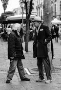 Two elderly people are engaged in conversation on a busy street. One is wearing a dark coat and a wide-brimmed hat, and the other is dressed in a bulky jacket. The scene is set in an urban area with a backdrop of trees and outdoor seating. There is a lively atmosphere, with several people in the background.